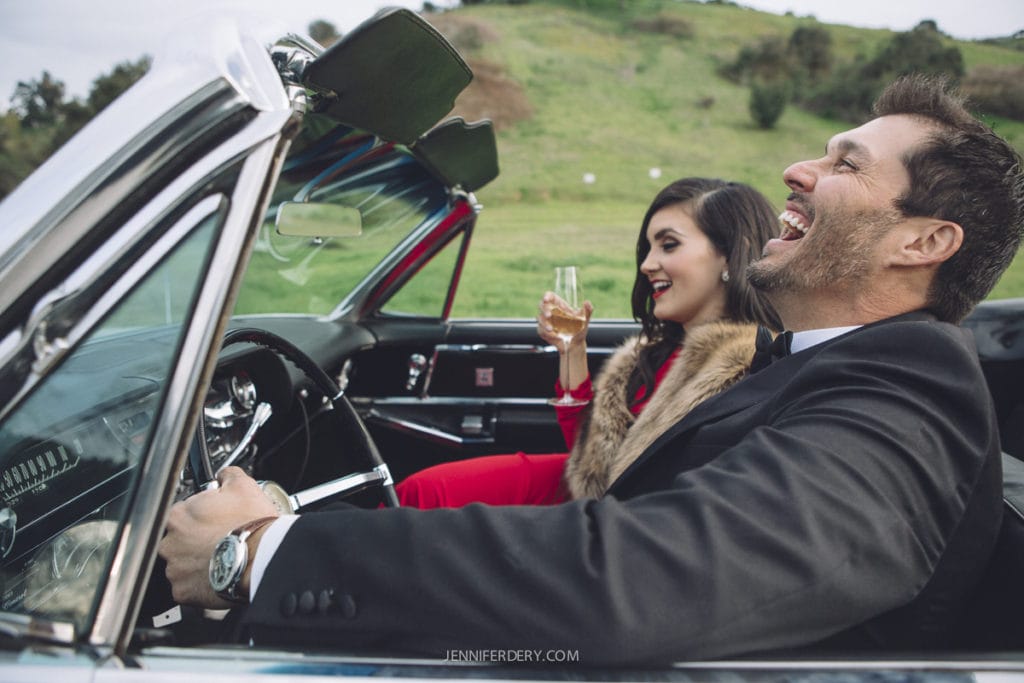 old-Hollywood glam-styled engagement session with a white vintage convertible car. shows black suite and red dress with fur stole