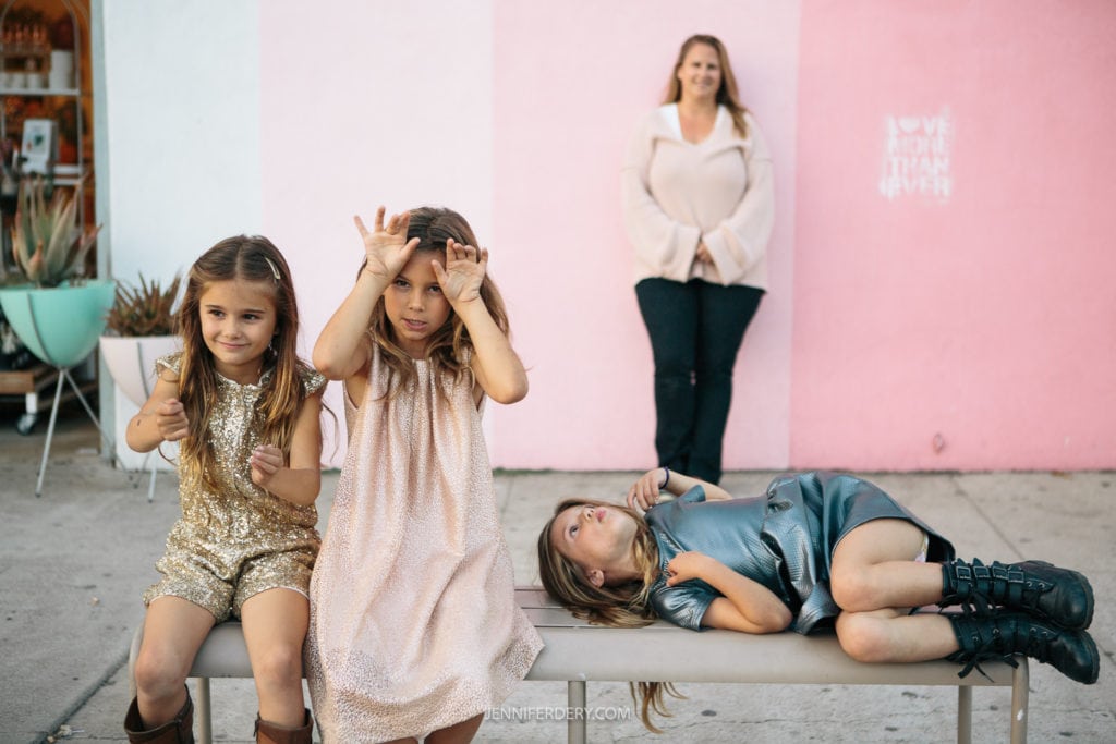 family of four in front of a multi-shaded pink wall in north park during their San Diego Family Portrait Session. A mom and 3 daughters are making goofy faces for the camera.