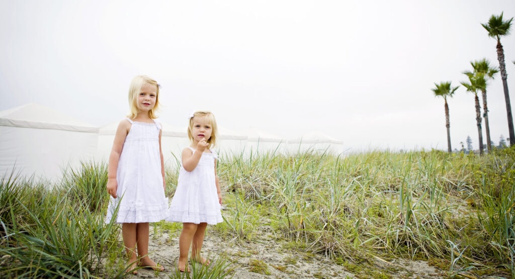 photo of two little kids dressed in white sun dresses on the beach in coronado standing in green beach grass