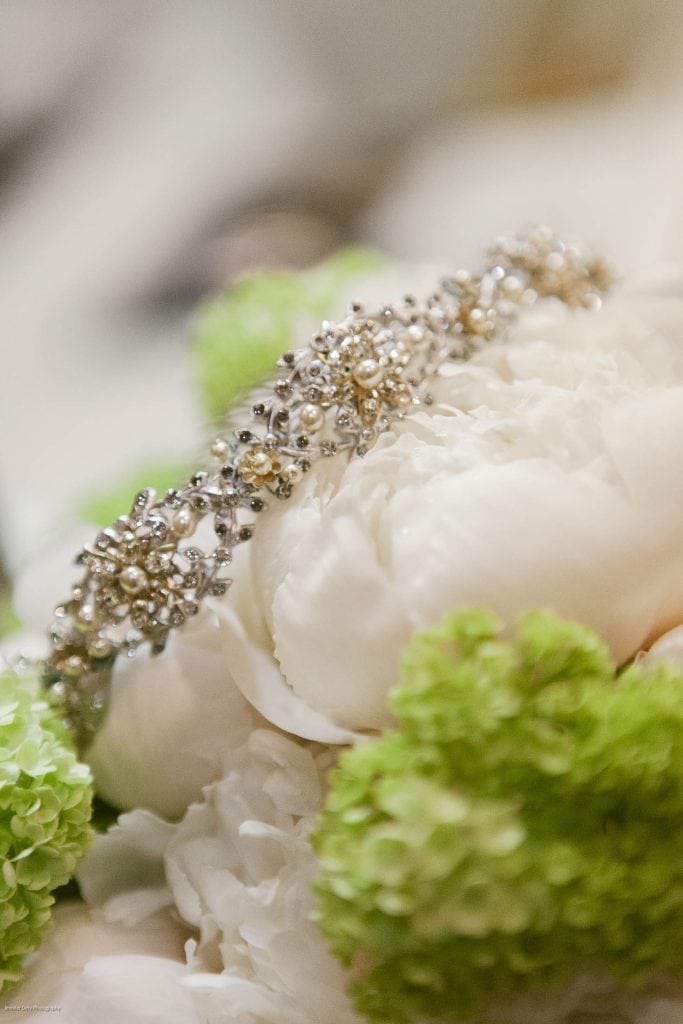 A delicate silver and pearl bridal headpiece rests on soft white peonies, surrounded by light green hydrangea blooms, creating an elegant and romantic scene.