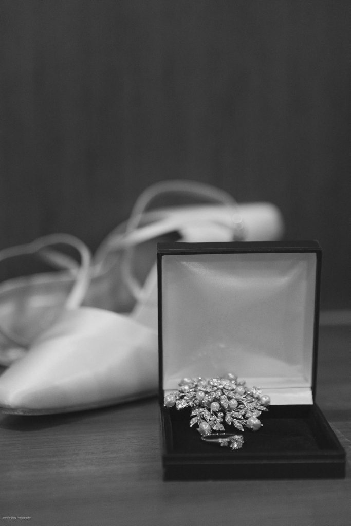 A jeweled brooch in an open velvet box sits on a wooden surface, with blurred white ballet flats and ribbon in the background. The image is in black and white.