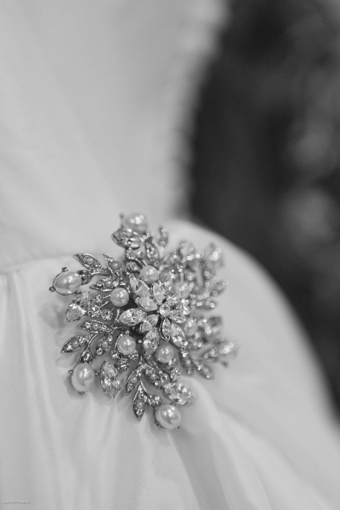 A close-up, black-and-white photo of an ornate brooch adorned with pearls and sparkling gemstones, fastened to a smooth, light-colored fabric, possibly part of a dress.