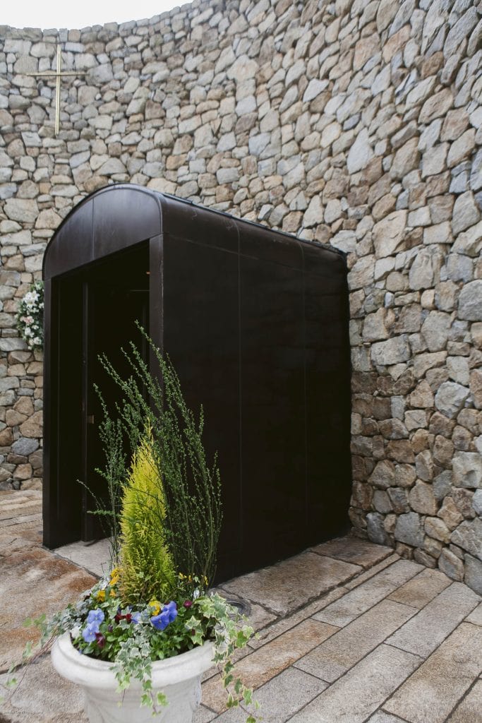 A small black structure stands against a high stone wall with a cross above it. In the foreground, there is a white planter with green plants and colorful flowers.