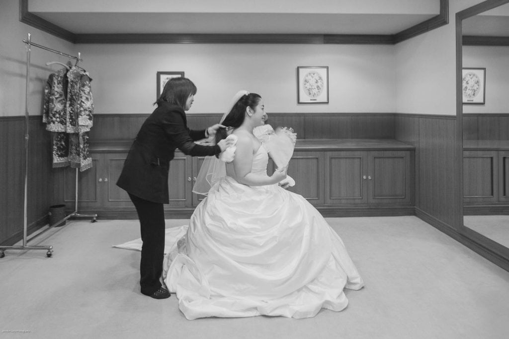 A woman in a wedding dress sits while another person helps arrange her hair in a fitting room. Wedding attire hangs on a rack, and the scene is reflected in a large wall mirror.