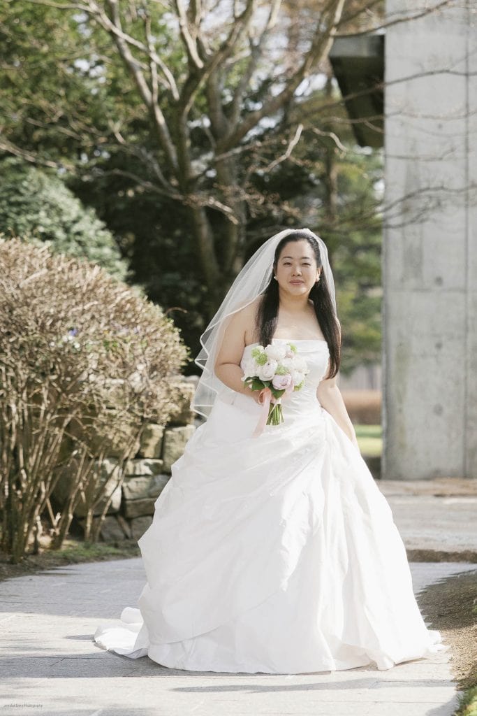 A bride in a white wedding dress and veil holds a bouquet of flowers while standing outdoors on a sunny day, surrounded by green bushes and trees.