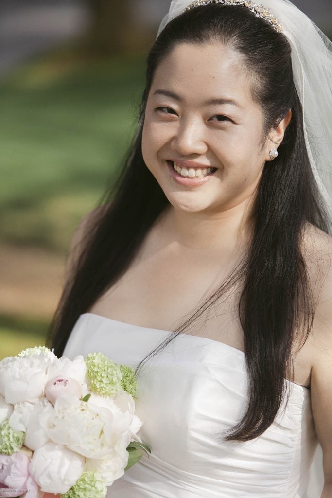A smiling bride with long dark hair, wearing a white strapless wedding dress and veil, holds a bouquet of white and light pink flowers outdoors on a sunny day.