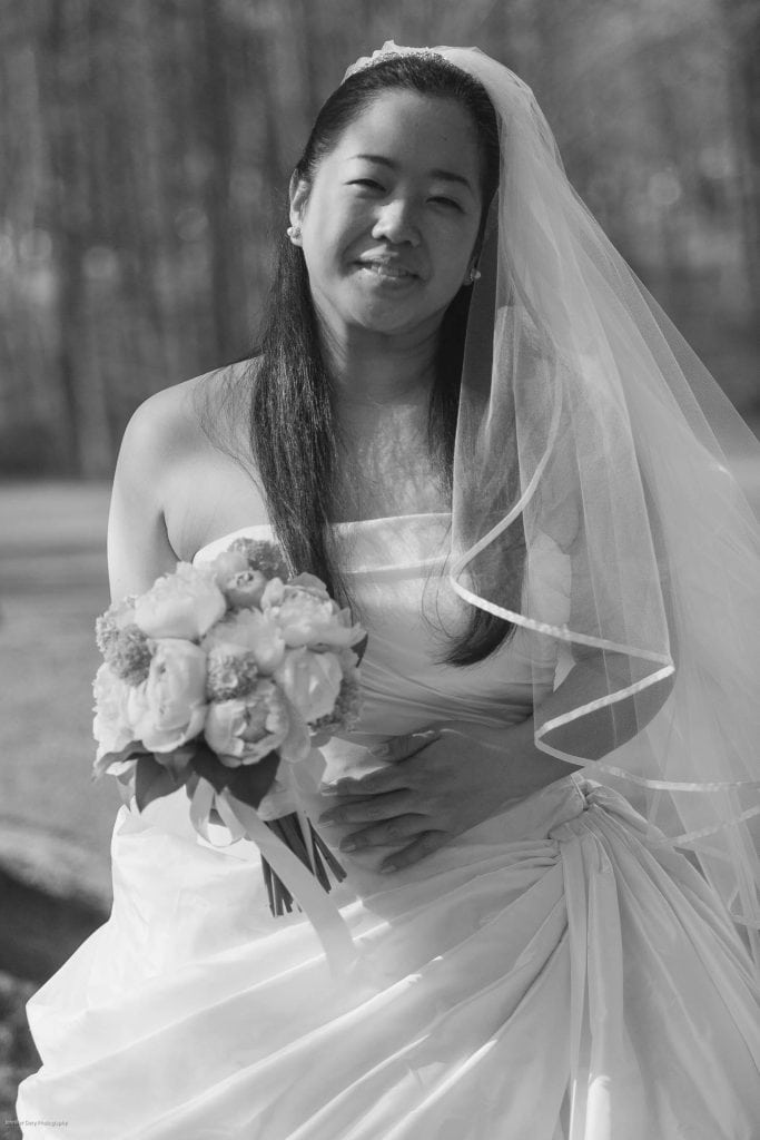 A bride in a strapless wedding dress and veil holds a bouquet of flowers, smiling softly outdoors with trees blurred in the background. The image is in black and white.