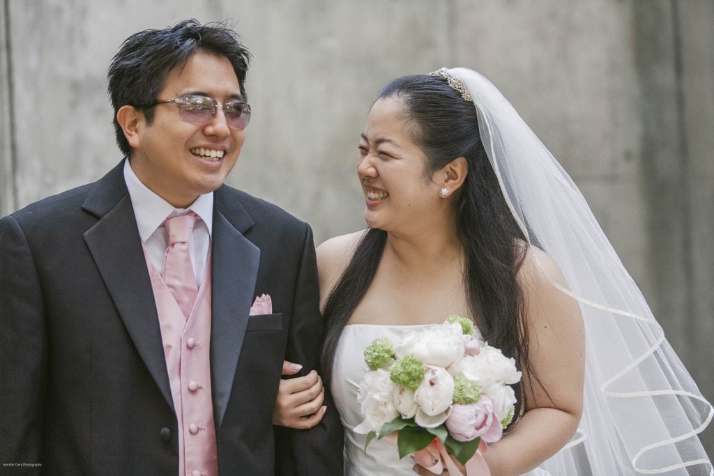 A joyful bride in a white dress and veil holds a bouquet of flowers, smiling and looking at a groom in a black suit, white shirt, and pink tie; they are walking arm in arm and laughing together.