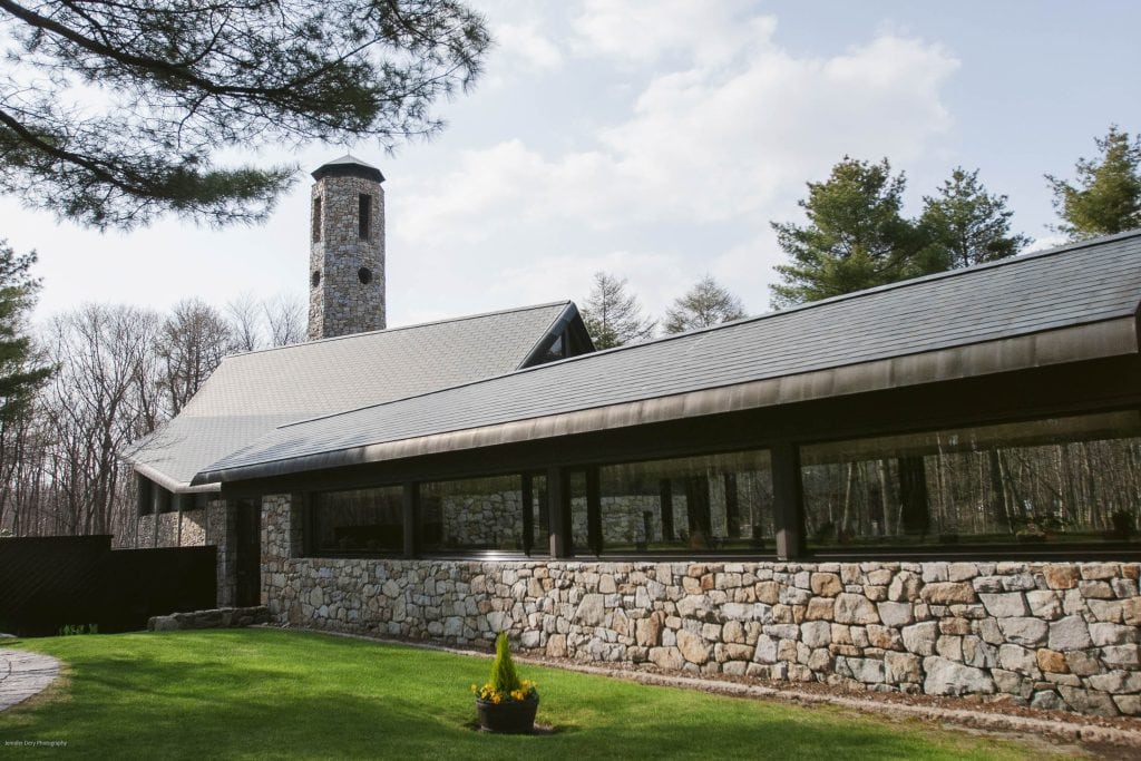 A stone building with a tall chimney, large windows, and a sloped roof is surrounded by green grass, trees, and a small planter with yellow flowers in the foreground.