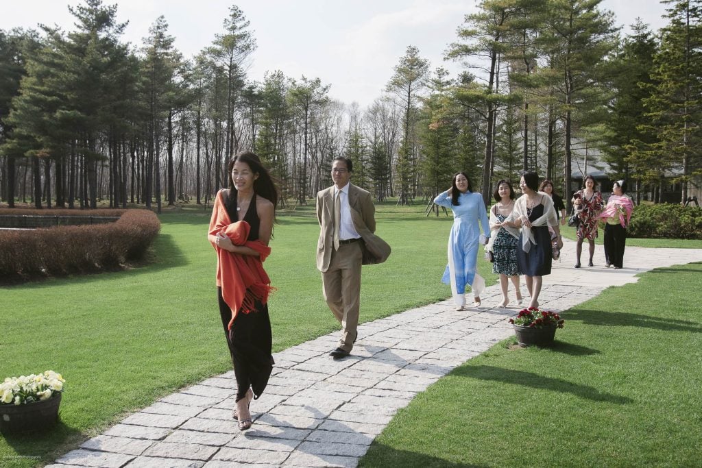 A group of people dressed in semi-formal attire walk along a stone path in a green park, with trees and a pond in the background. A woman in a black dress and orange shawl leads the group, smiling.