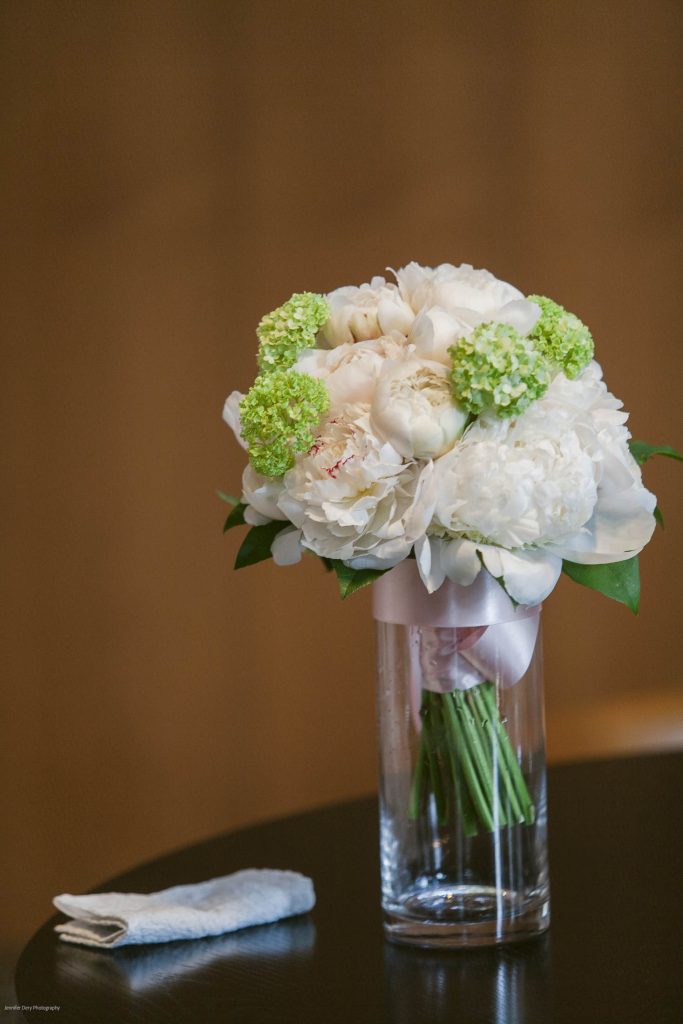 A bouquet of white and light green flowers in a clear glass vase with a pink ribbon sits on a dark table next to a folded white handkerchief, against a soft brown background.