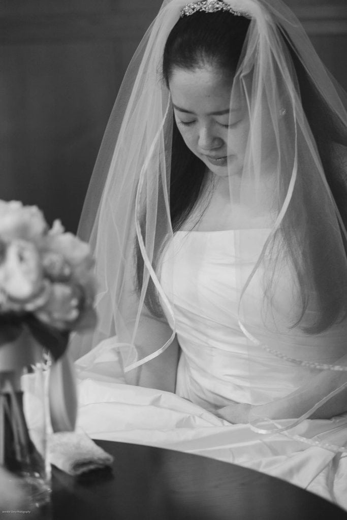 A bride in a white wedding dress and veil sits, looking down peacefully. A bouquet of flowers in a vase is visible in the foreground. The image is in black and white.
