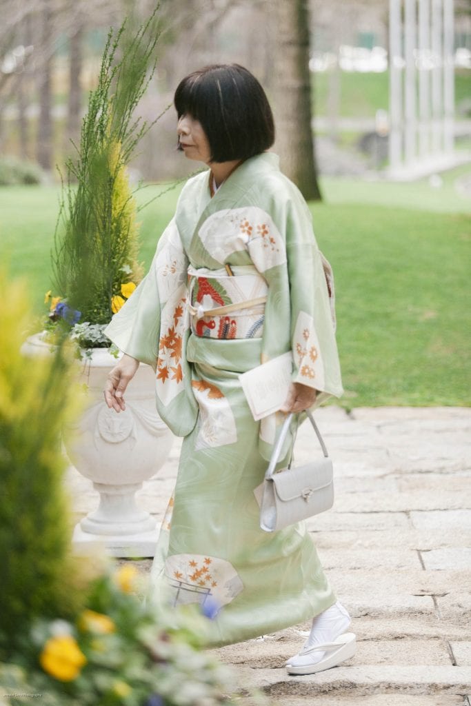 A woman in a light green kimono with floral patterns walks on a stone path, holding a gray handbag and a folded paper. She is outdoors, with greenery, trees, and potted plants in the background.