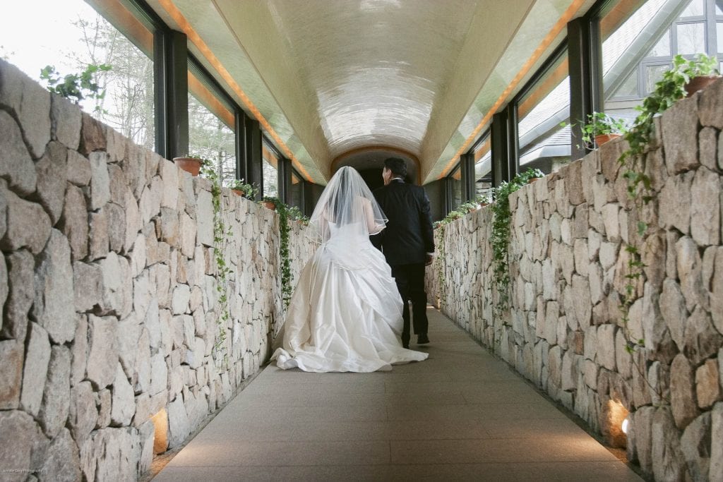 A bride in a white gown and veil walks hand in hand with a groom in a black suit down a stone hallway lined with green plants and soft lights.