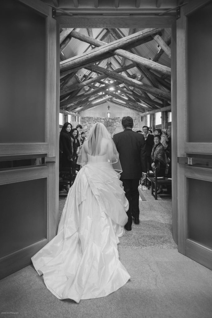 A bride in a white gown and veil walks down the aisle with a man inside a chapel, as guests seated on either side watch. The church features wooden beams and a cross at the front.