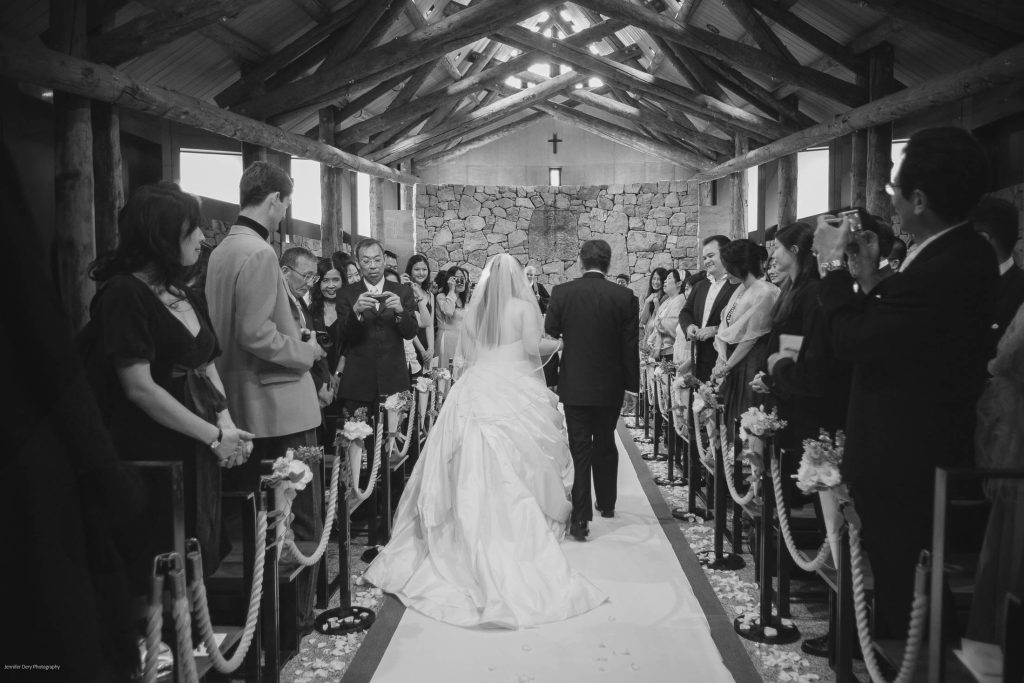 A bride in a white gown walks down the aisle with a man, likely her father, in a rustic chapel as guests stand, watch, and take photos. The setting is decorated with flowers and wooden beams overhead.