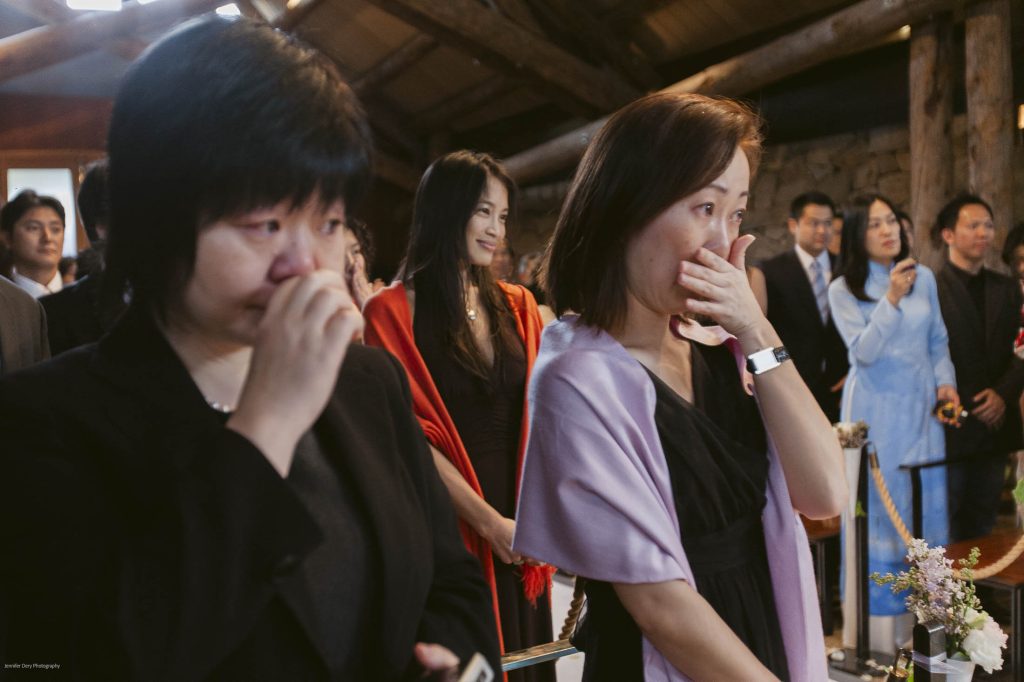 Two women in the foreground wipe tears from their faces at an indoor event, while other well-dressed guests stand behind them, some smiling and others watching solemnly. The setting appears warm and rustic with wooden beams.
