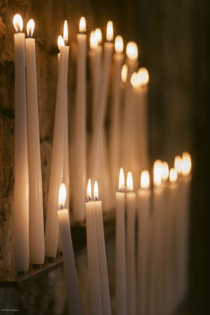 Rows of tall, white candles are lit and placed on metal holders against a stone wall, creating a warm, glowing atmosphere in a dimly lit space.