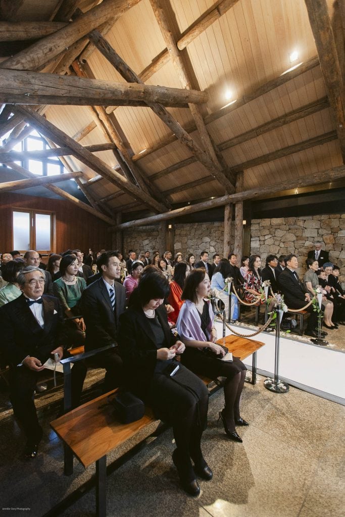 Guests seated in a rustic, wooden-beamed chapel with stone walls, attentively watching a ceremony taking place at the front. Sunlight filters through the windows, creating a warm and inviting atmosphere.