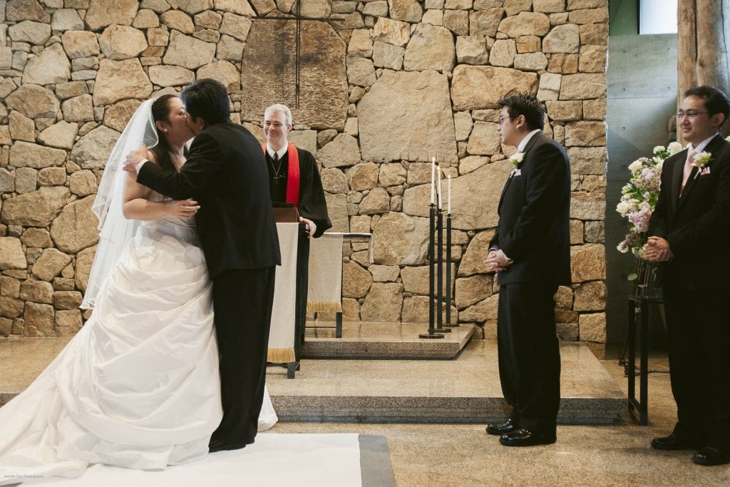 A bride is kissed on the cheek by an older man, likely her father, during a wedding ceremony. The groom and another man stand to the right, while an officiant smiles in the background against a stone wall.