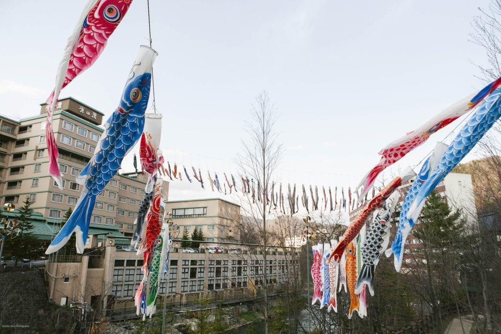 Colorful koi-shaped windsocks (koinobori) are strung across an outdoor area near buildings and trees, fluttering in the breeze under a clear sky. The scene appears festive and vibrant.