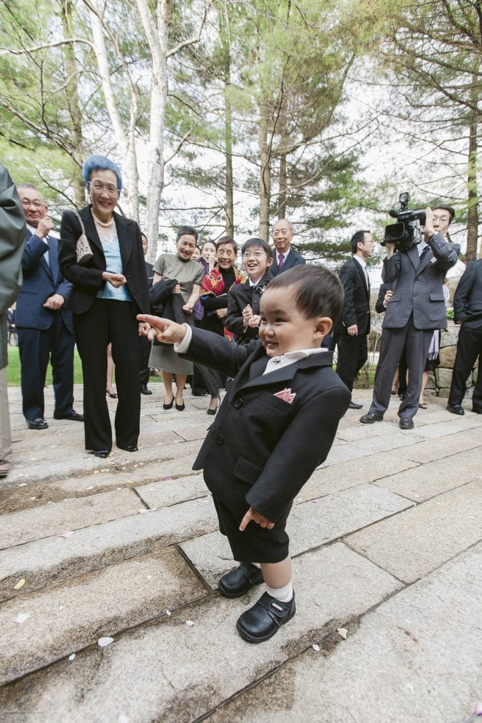 A young boy in a black suit smiles and points while standing on stone steps outdoors. Adults dressed formally stand behind him, some laughing, with trees and greenery in the background.