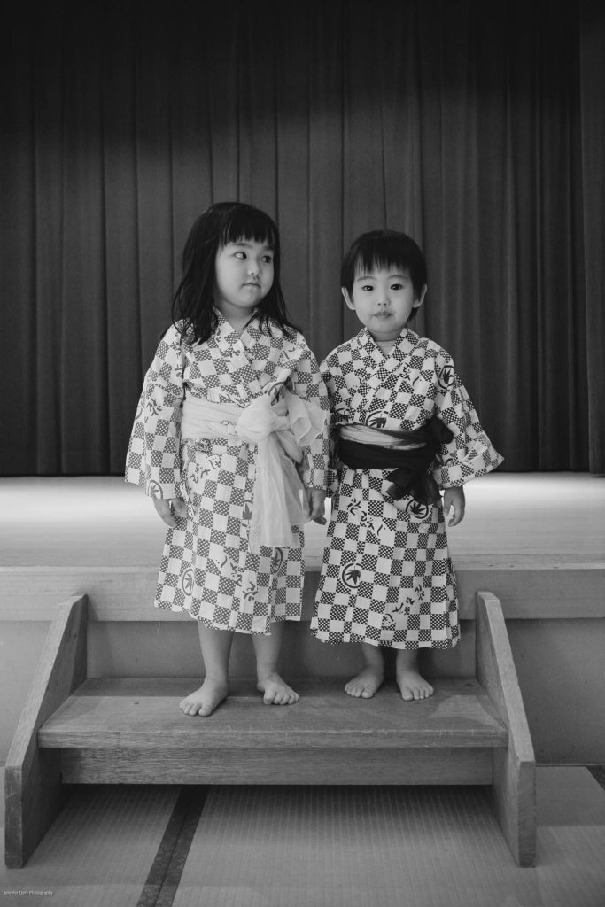 Two young children wearing traditional patterned robes stand barefoot on a small wooden platform in front of a stage with curtains. The child on the left looks to the side, while the child on the right looks at the camera.