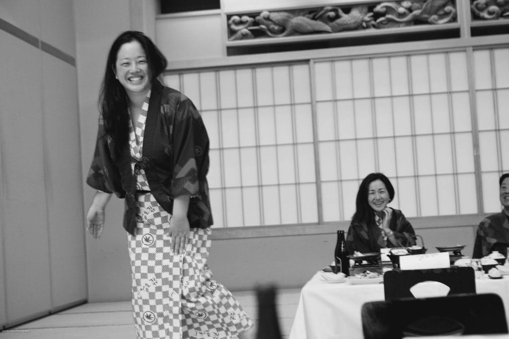 A woman in a patterned yukata smiles and walks past a low table while two people sitting at the table smile and clap in a traditional Japanese-style room. The photo is in black and white.