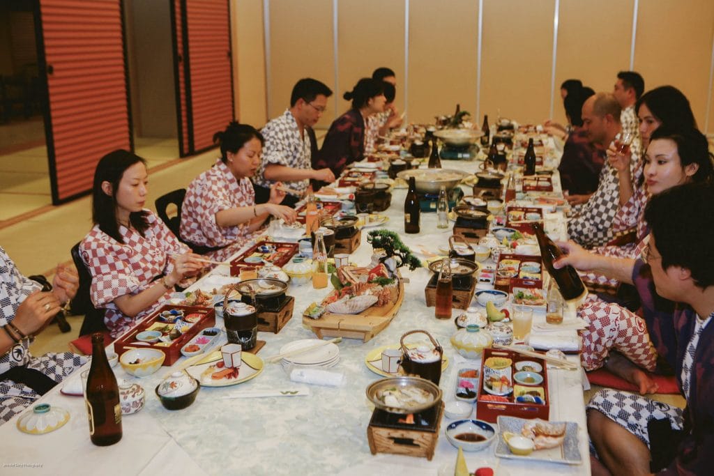 A group of people wearing traditional Japanese attire sit around a long table, enjoying a meal with various dishes, drinks, and bento boxes in a brightly lit room.