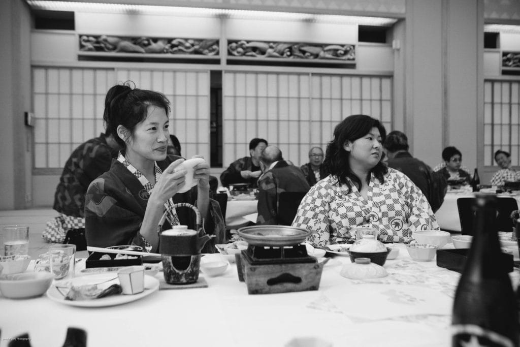 Two women in traditional Japanese robes sit at a table set with various dishes and drinks in a tatami room. The background shows other people dining and sliding shoji doors. The scene is captured in black and white.