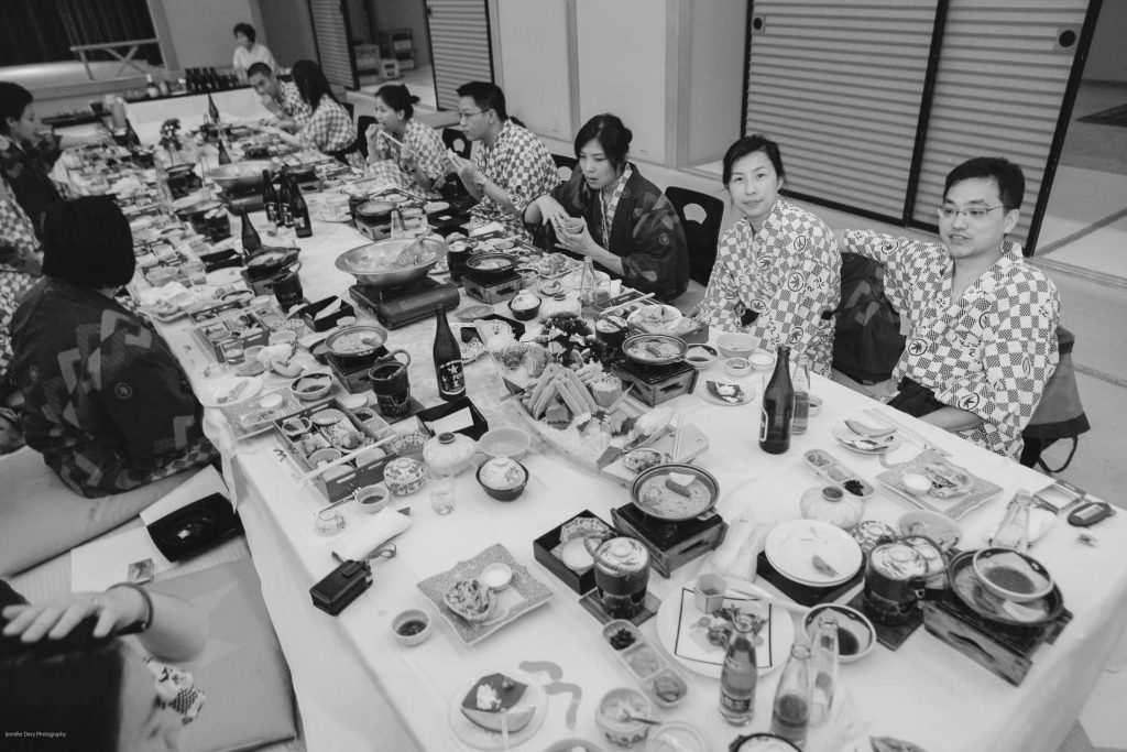 A group of people wearing patterned robes sit around a long table filled with various Japanese dishes, drinks, and hot pots, sharing a meal in a festive, indoor setting.