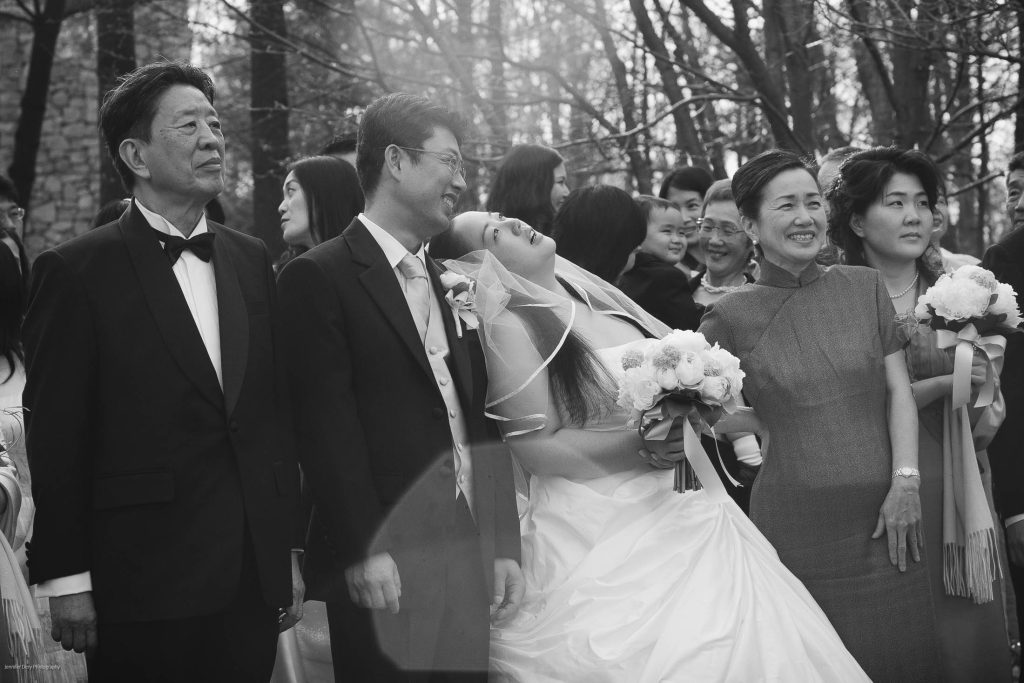 A bride laughs joyfully, leaning back beside the groom and family members, all dressed formally, holding bouquets at an outdoor wedding ceremony. The mood is joyful and celebratory.
