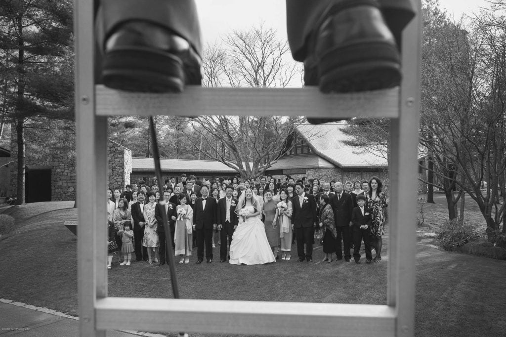 A large wedding group poses outdoors, facing the camera. The photo is taken from behind a ladder, with a pair of shoes and legs visible at the top, framing the group below. The setting includes trees and buildings.