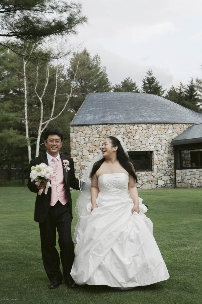 A bride and groom, both smiling, walk hand in hand on a green lawn in front of a stone building. The bride wears a white gown and the groom wears a black suit with a pink vest and holds a bouquet.