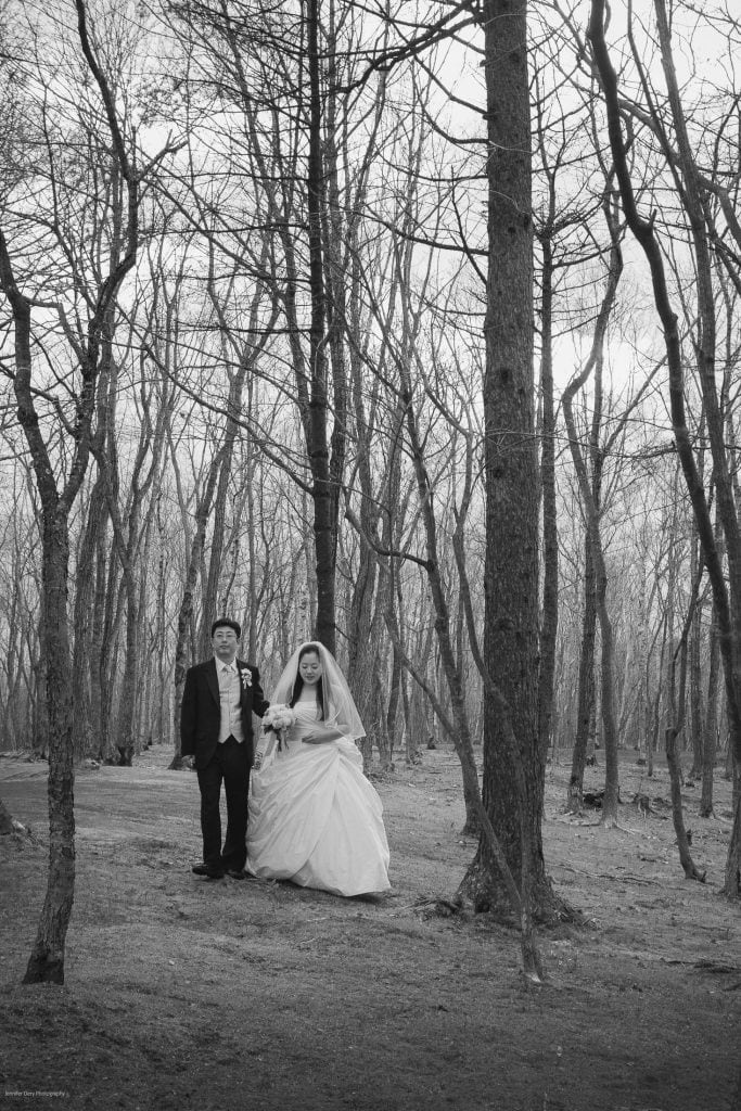 A bride in a white gown and a groom in a suit walk together through a forest of bare trees, holding hands, on a clear day. The scene is in black and white.