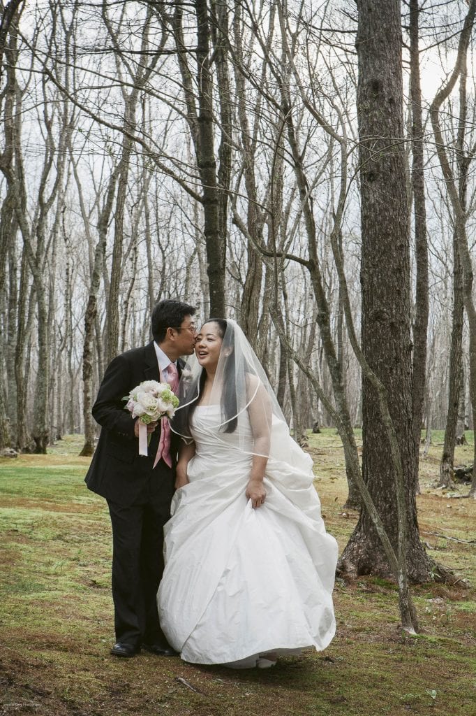 A groom in a dark suit kisses his bride, who is wearing a white wedding dress and veil, as they stand together in a forest with bare trees. The bride holds a bouquet and both are smiling.