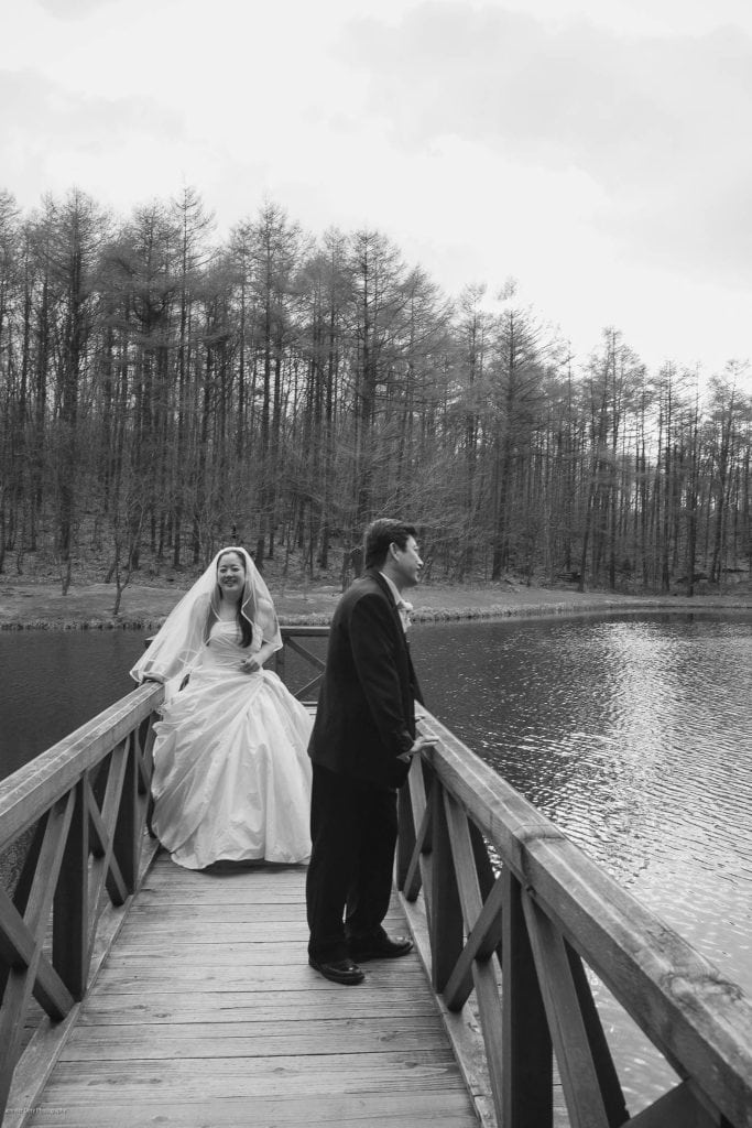 A bride in a gown and veil stands on a wooden bridge over a lake, while a man in a suit stands near her, with leafless trees in the background. The scene is in black and white.