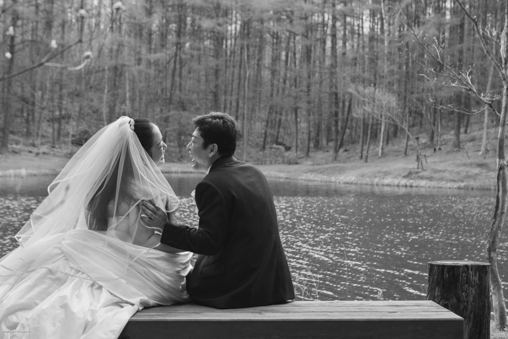 A bride and groom sit closely on a bench by a lake, facing each other and smiling. The bride wears a veil and gown; the groom is in a suit. Trees surround the calm water in the peaceful, outdoor setting.