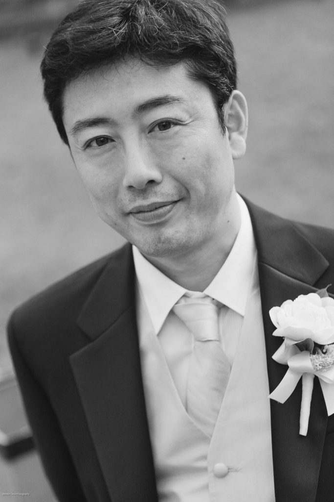 A man in a suit with a boutonnière on his lapel smiles gently at the camera. The image is black and white, and the background is softly blurred.