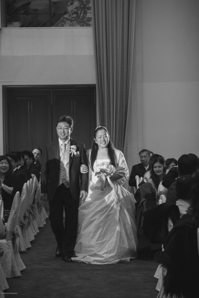 A bride and groom walk arm-in-arm down an aisle, smiling, surrounded by seated guests who are clapping and watching them. The setting appears to be an indoor wedding ceremony.