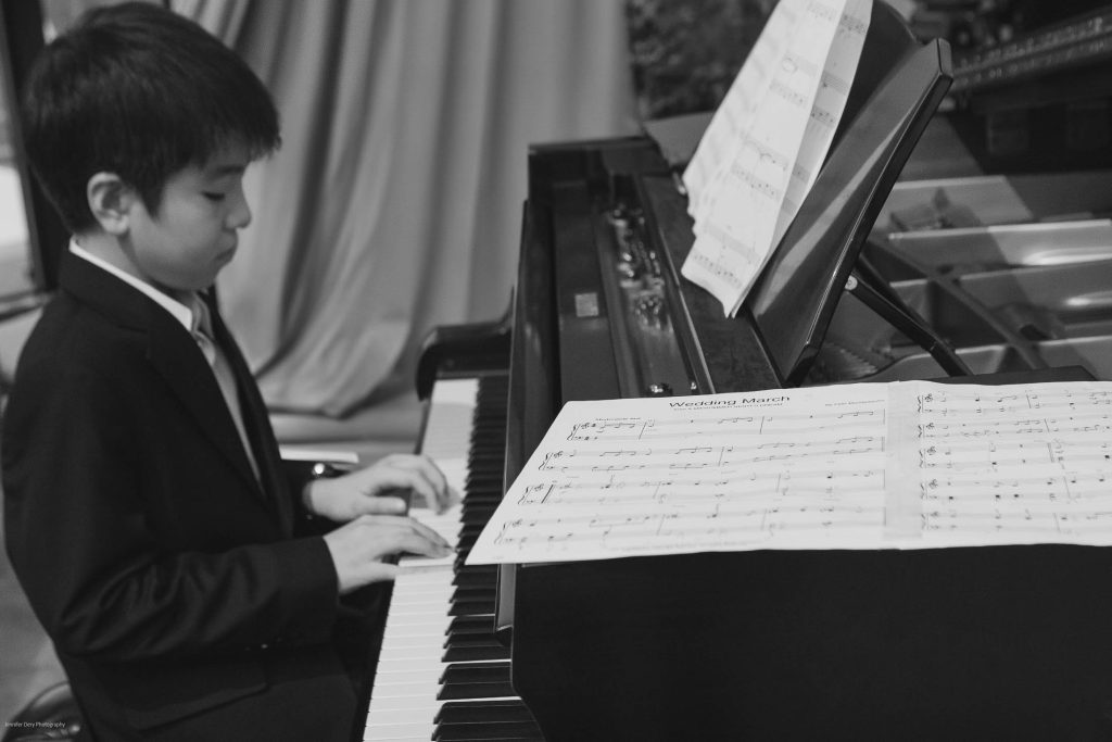 A young boy in a suit plays the piano, reading sheet music placed on the instrument. The image is in black and white, and draped curtains are visible in the background.
