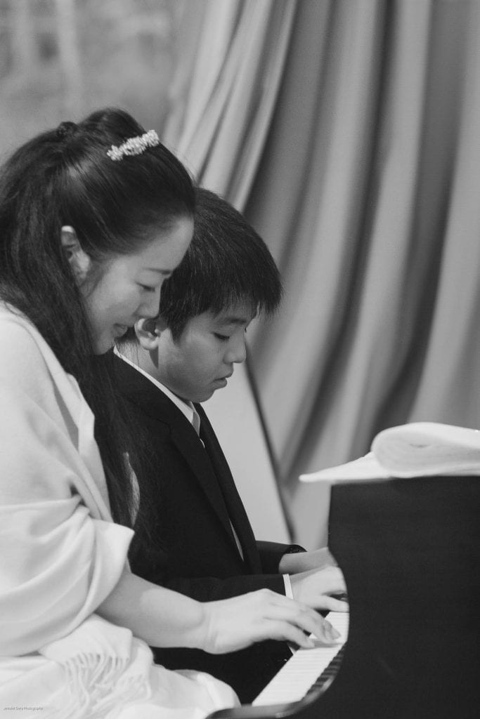 A woman and a boy play the piano together, seated side by side. They both look focused on the keys. The image is in black and white, with a curtain in the background.
