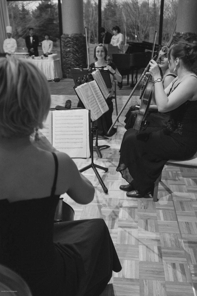 A black-and-white photo of a string quartet of women in formal dresses performing indoors, with music stands in front of them. In the background, chefs and guests stand near a grand piano and large windows.
