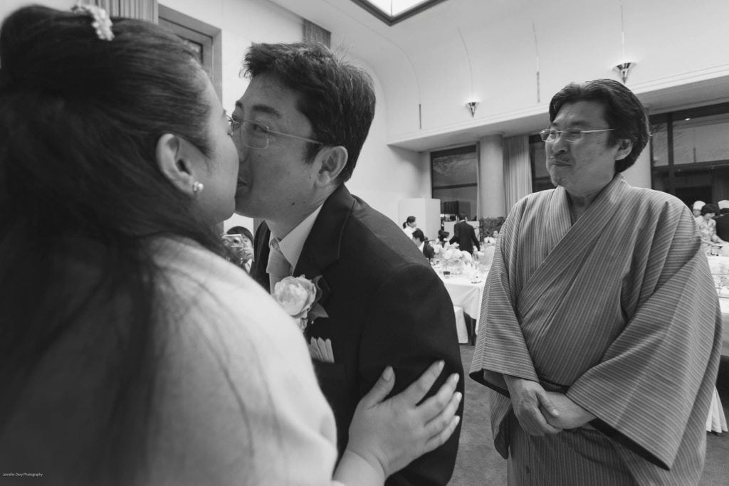 A bride and groom share a kiss at their wedding reception while a smiling man in traditional Japanese attire stands nearby, watching them. Guests are seated at tables in the background.