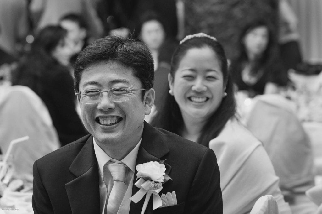 A man in a suit and tie with a boutonnière and a woman in a white dress sit side by side, both smiling and laughing, surrounded by guests at a formal event.