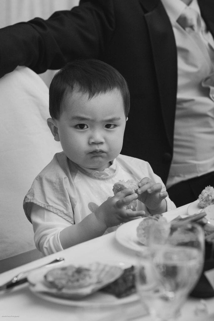 A young child with a serious expression sits at a table, holding food in both hands, with plates of food and a glass in front of them. An adult in a suit sits beside the child. The image is in black and white.