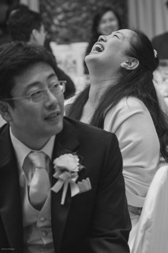 A woman in a white dress laughs heartily while sitting beside a man in a suit with a boutonniere at a formal event, possibly a wedding. Other guests are visible in the blurred background.