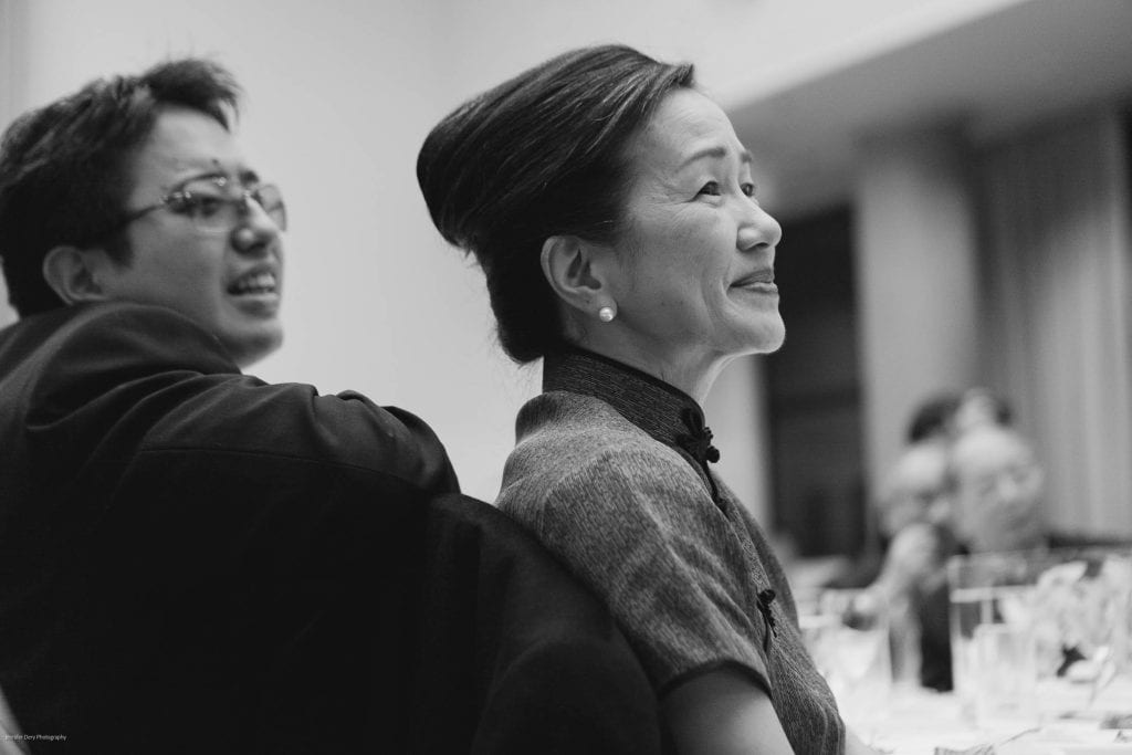 A man and a woman sit closely together at a formal event, both smiling and looking toward the front. The woman wears a traditional dress and pearl earrings. The image is in black and white.