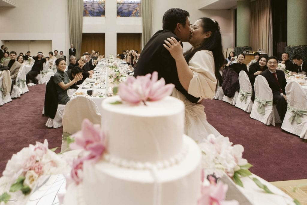 A bride and groom kiss at their wedding reception, standing behind a white, flower-decorated cake. Guests seated at long tables watch and smile, with some clapping and taking photos. The scene is joyful and festive.