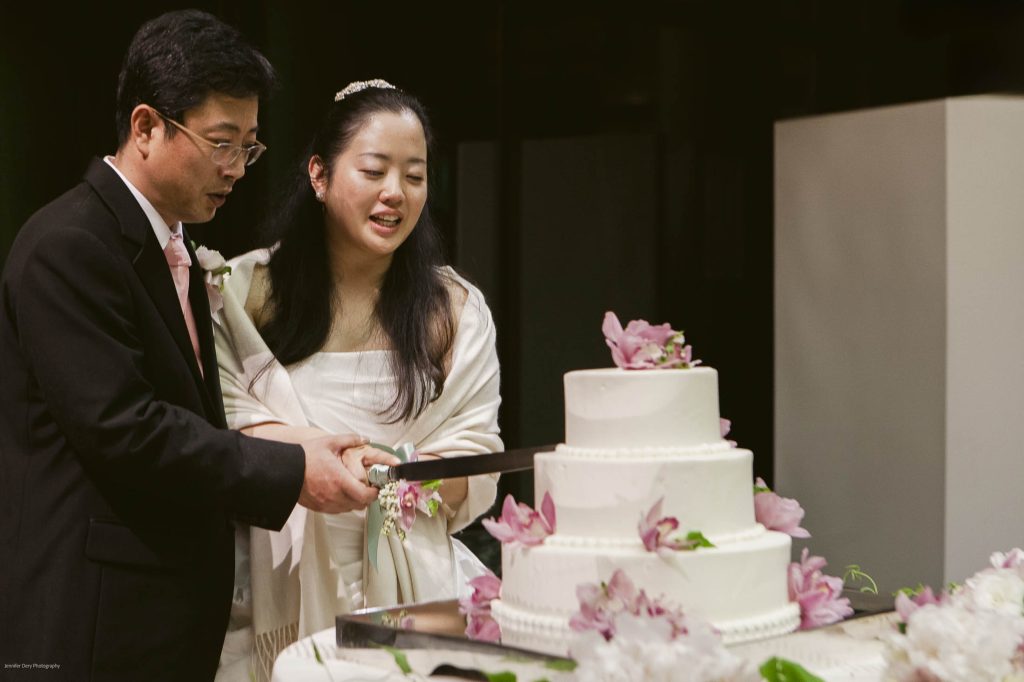 A bride and groom stand together, smiling as they cut a white, three-tiered wedding cake decorated with pink flowers. The groom wears a black suit and the bride is in a white dress with a shawl.